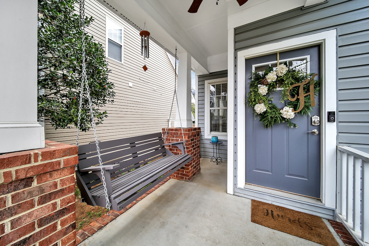 A welcoming porch area features a wooden swing adorned with cushions, set against a brick wall. A decorative wreath hangs on the gray front door, complemented by a small blue decorative piece nearby. Natural light flows into the space, enhancing its inviting feel.