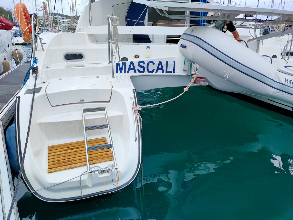 The image displays the bow of a catamaran named 'Mascali', securely docked. A small motorboat is tied alongside, with wooden steps leading onto the deck. The surrounding water is calm and has a rich turquoise hue, with other boats visible in the background.