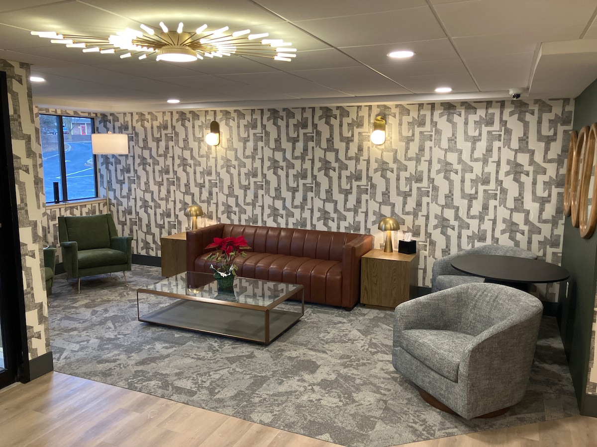 A modern lobby area features a brown leather sofa set against a patterned wall, complemented by a glass coffee table. Two accent chairs in green and grey provide additional seating, while warm lighting is offered by stylish wall sconces and table lamps.
