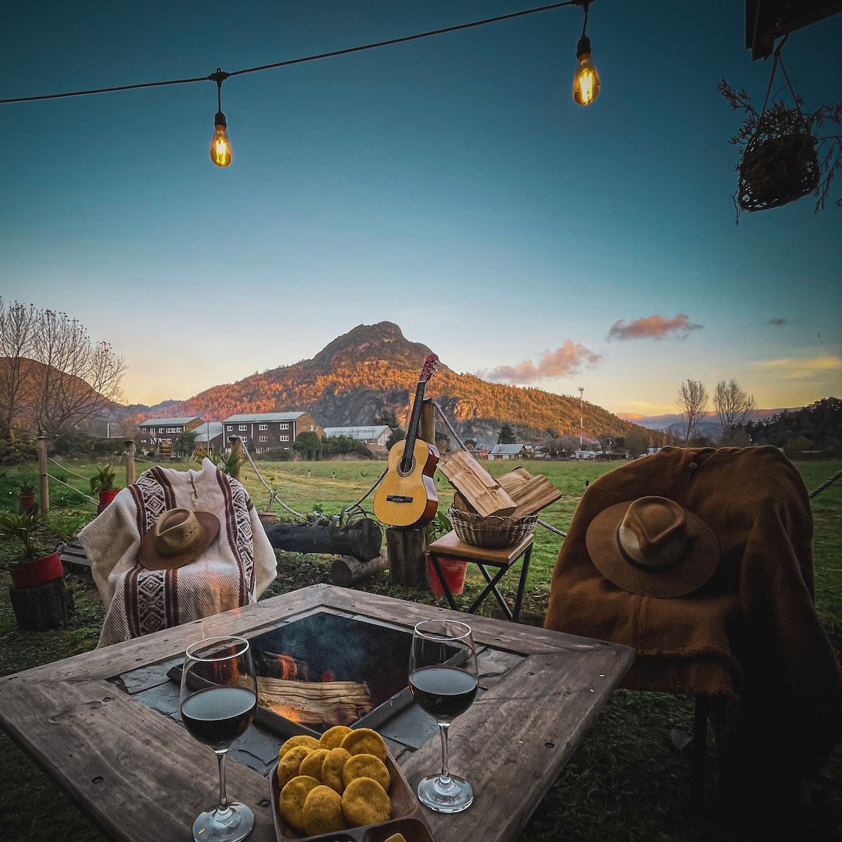 A cozy outdoor seating area is presented, featuring a fire pit surrounded by three chairs. Clear glasses of wine sit atop a wooden table, accompanied by a plate of snacks. A view of a mountainous landscape is visible in the background, under a twilight sky.