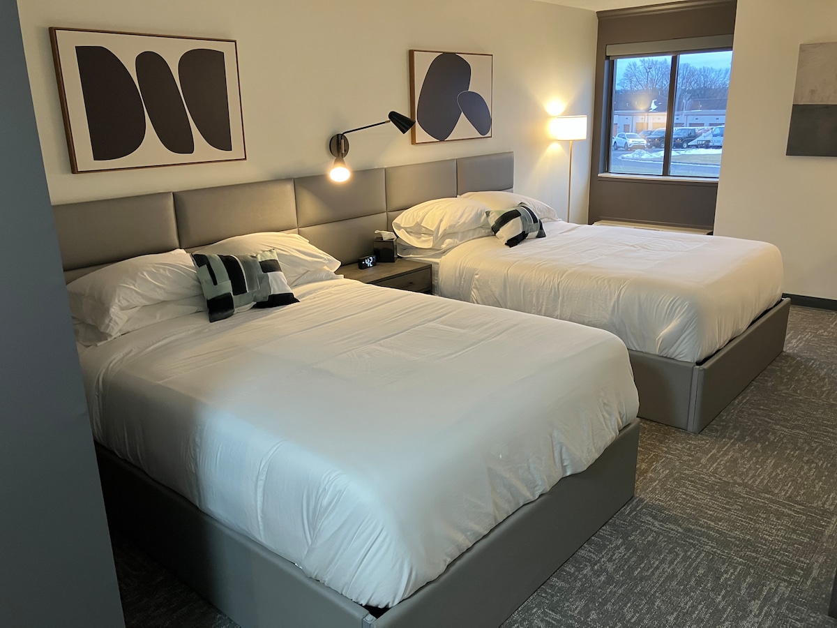 The room features two spacious beds with white bedding, complemented by stylish wall art. A bedside table with a lamp stands between the beds. Natural light enters through a large window, offering views of the surroundings.