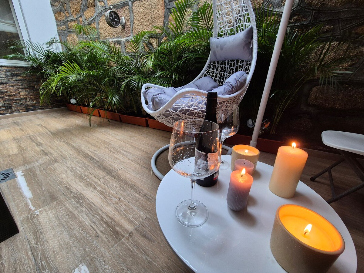 A cozy outdoor space features a white hanging chair surrounded by lush greenery. A round table holds a wine glass, a dark bottle, and several candles in varied heights, providing a relaxed atmosphere for enjoying a serene moment.