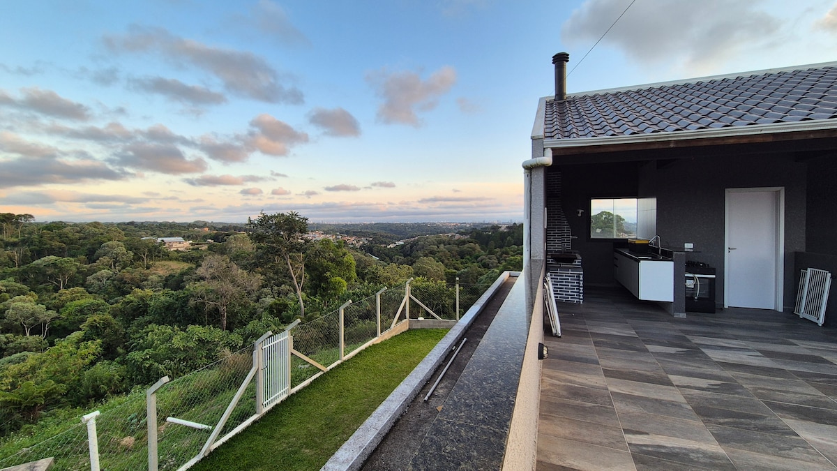 A spacious outdoor area offers a stunning view of rolling hills and greenery. A modern kitchen space is partially visible, featuring sleek counters. Surrounding fencing and well-maintained grass frame the setting, while the sky transitions from soft blue to warm hues of sunset.
