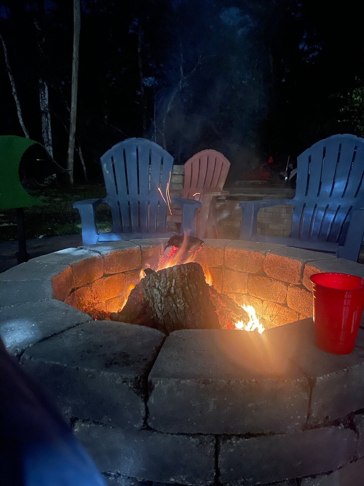 A circular stone fire pit is illuminated by a crackling fire, with warm orange and yellow flames visible. Surrounding the fire are several adirondack chairs in shades of blue and pink. A red cup sits nearby, adding a pop of color to the tranquil outdoor setting.
