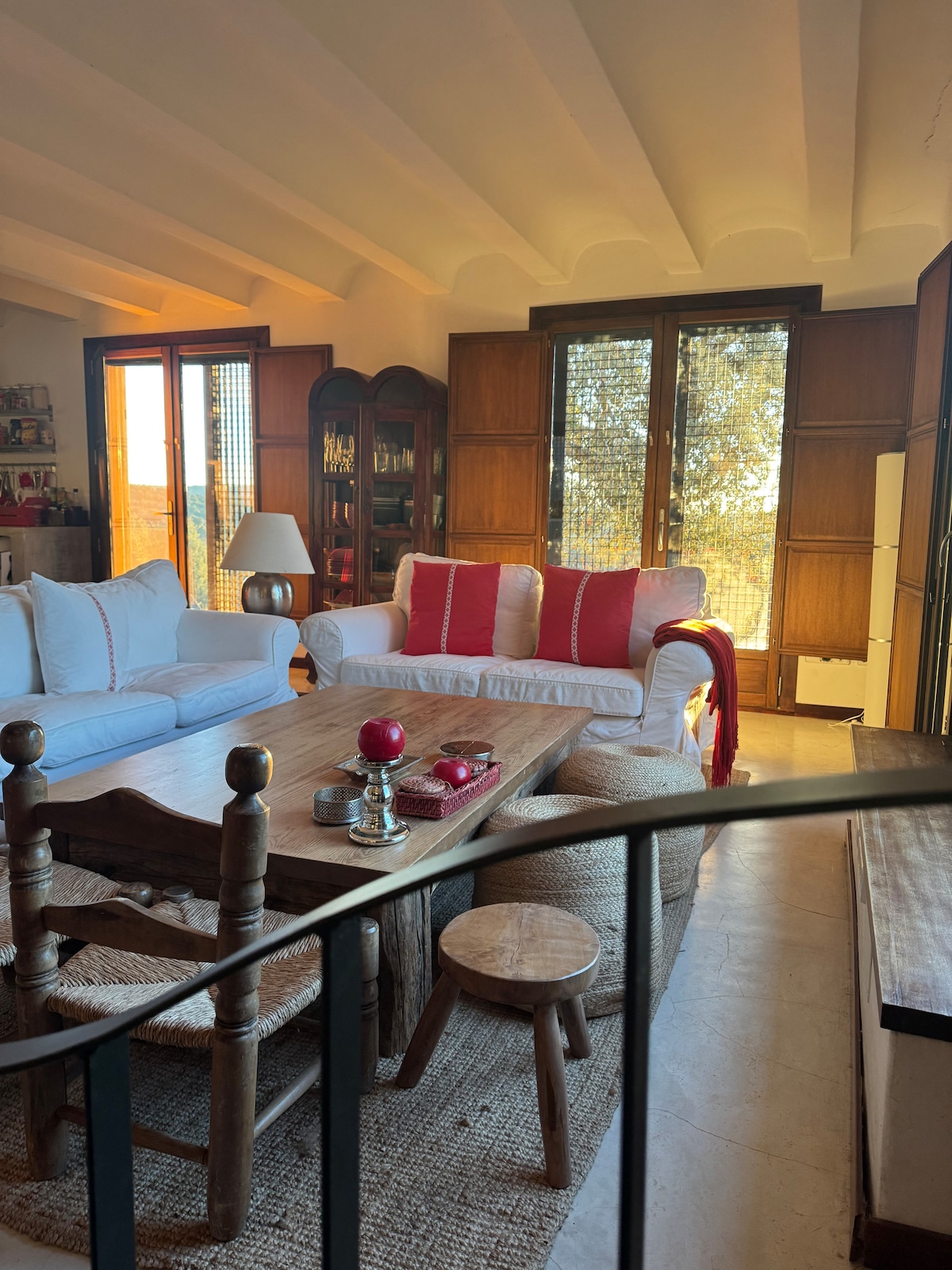 A bright living area features a light-colored sofa adorned with red accent pillows. A wooden coffee table is centered among woven stools, and warm light filters through large windows, revealing views of the surrounding nature. A welcoming atmosphere is created by natural textures and earthy tones.