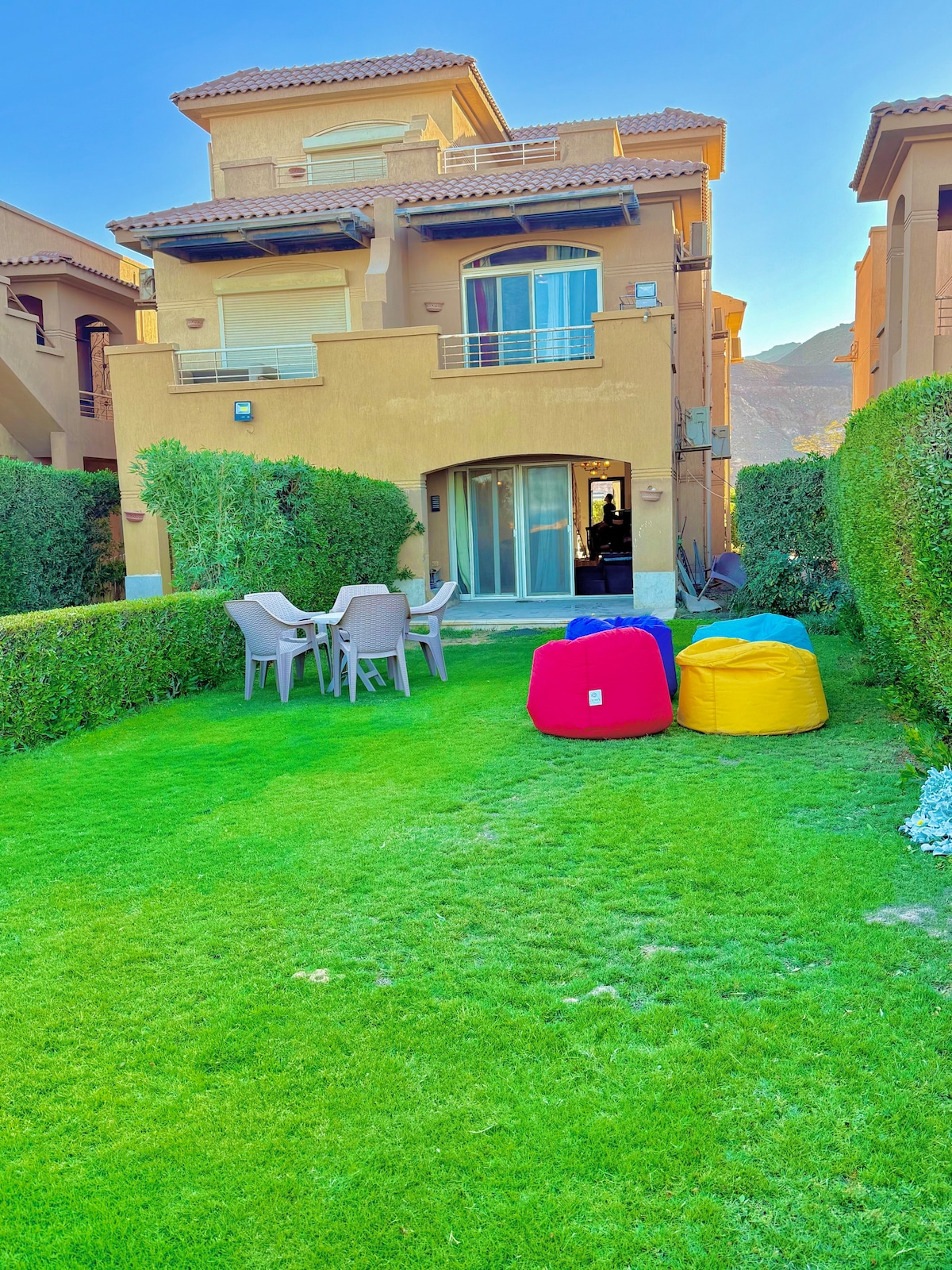 A well-maintained garden area features a neatly manicured lawn surrounded by green hedges. Colorful bean bag chairs are placed invitingly alongside a set of outdoor dining furniture, consisting of a round table with accompanying chairs, creating a relaxed outdoor space.
