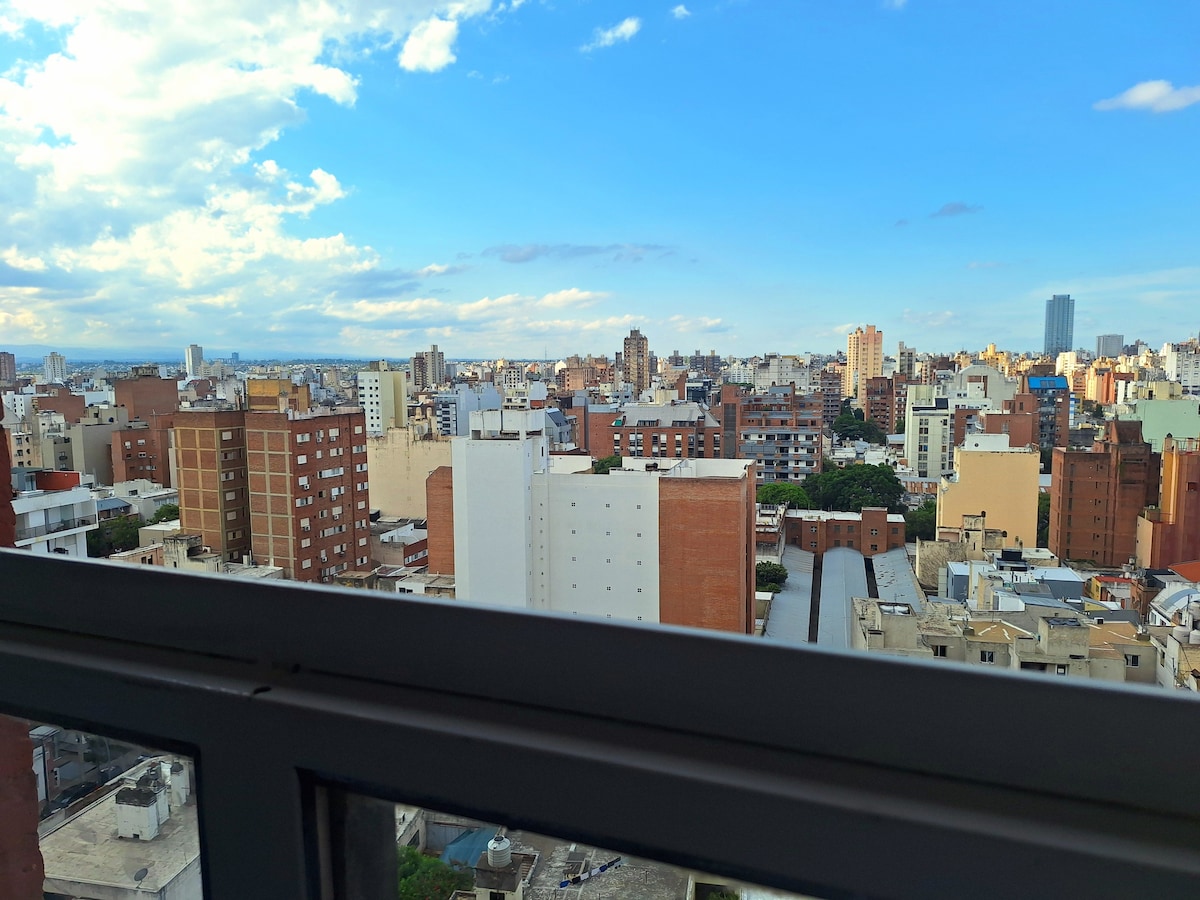 A panoramic view of the cityscape showcases a mix of residential and commercial buildings, interspersed with green rooftops. The skyline features various architectural styles under a bright blue sky with soft clouds, conveying an urban atmosphere.