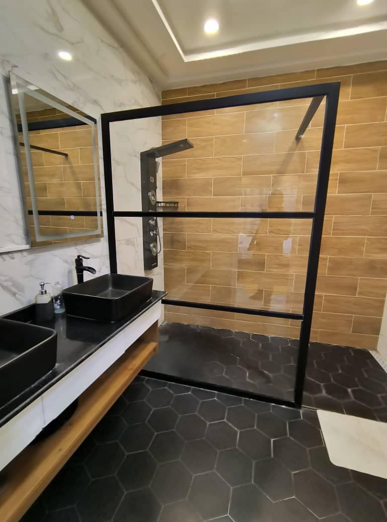 The bathroom features a spacious shower area enclosed by sleek black glass. A modern sink sits on a wooden vanity, complemented by a large wall mirror. Floor tiles in a hexagonal pattern add a contemporary touch to the design.
