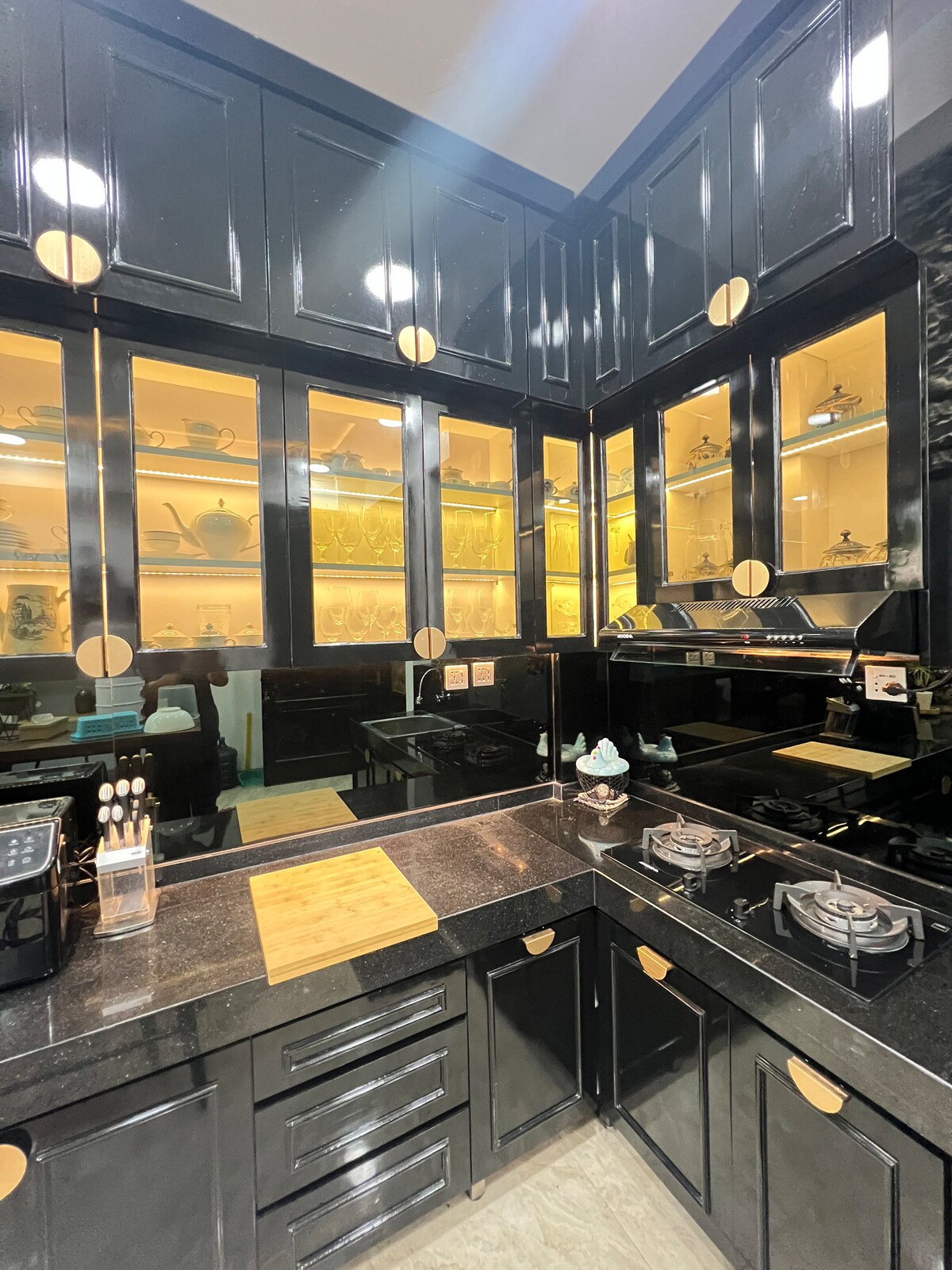 The kitchen features sleek black cabinetry with elegant gold handles and glass-fronted upper cabinets, showcasing various glassware. A dark countertop complements the modern appliances, including a gas stove. Warm lighting enhances the sophisticated atmosphere.