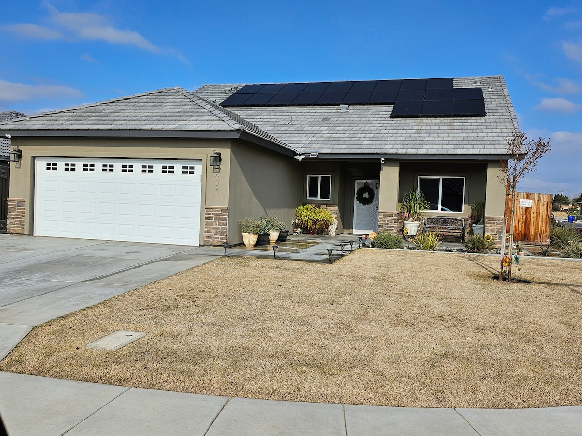 A one-story home features solar panels on the roof and a sturdy stone facade. The driveway accommodates multiple vehicles, while a manicured lawn offers a tidy appearance. Potted plants enhance the front porch area, contributing to the home's overall inviting presence.