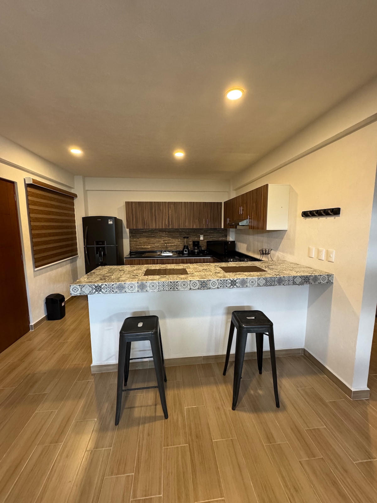 A modern kitchen is displayed with dark wooden cabinets and a decorative tiled countertop. Sleek black stools are positioned at the counter, and modern appliances, including a refrigerator, are visible. The space is well-lit, creating an inviting atmosphere for meal preparation.