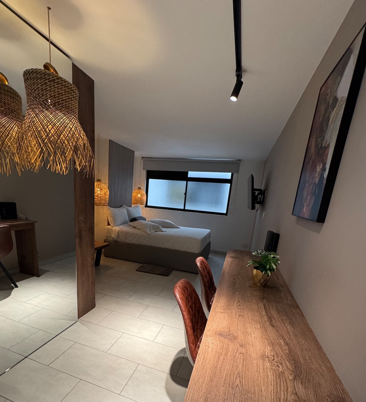A modern room features a spacious bed, complemented by natural light filtering through the window. Two woven light fixtures hang above a sleek wooden desk with two chairs, providing a functional workspace. A low partition separates the sleeping area from the entrance.