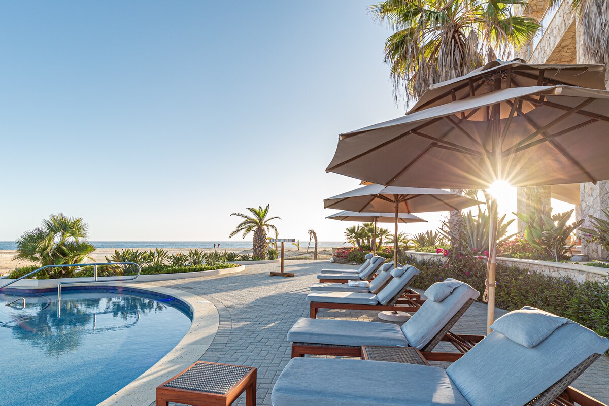 Comfortable lounge chairs with shades are arranged near a serene pool overlooking the beach. Lush greenery and palm trees surround the area, with sunlight glinting off the water's surface, creating a relaxing atmosphere.