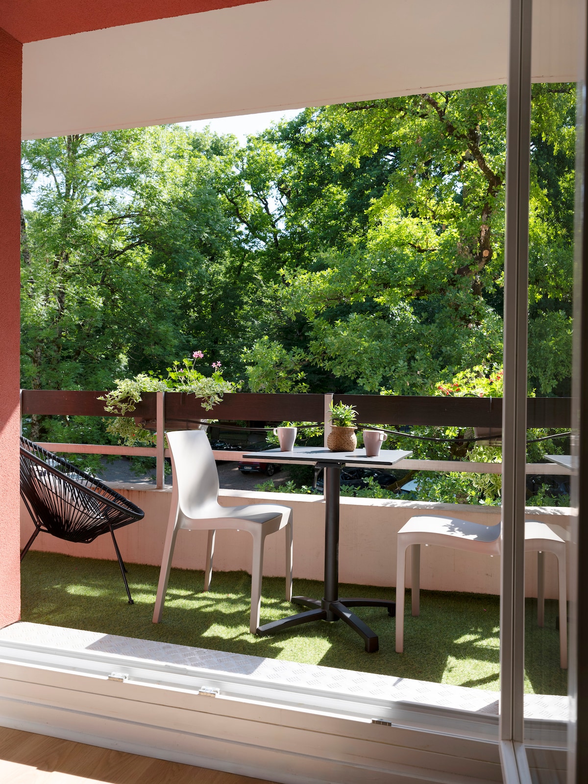 A balcony is presented with modern seating, including a small round table and two chairs. Potted plants enhance the natural setting, with a backdrop of lush green trees visible from the space.