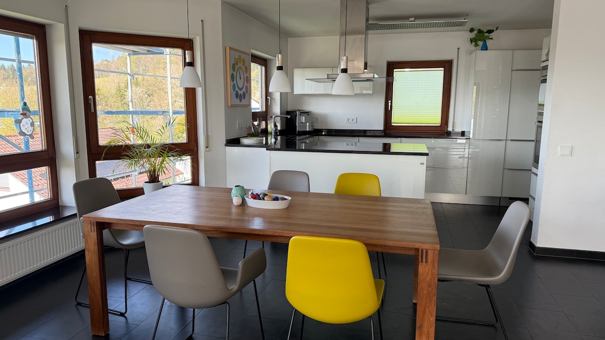 A spacious kitchen area is highlighted by a large wooden dining table surrounded by colorful chairs. Bright natural light filters through the expansive windows, creating a warm environment. The modern kitchen features sleek cabinetry and appliances, with plants adding a touch of greenery.