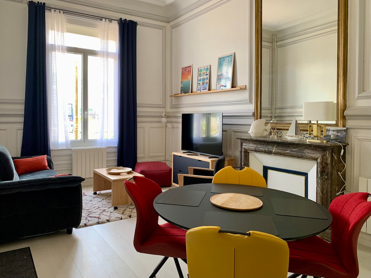 A bright living area features large windows dressed in light curtains, allowing natural light to fill the space. Comfortable seating is arranged around a coffee table. A sleek TV stand and a fireplace with marble accents are visible, complemented by a dining table surrounded by colorful chairs.