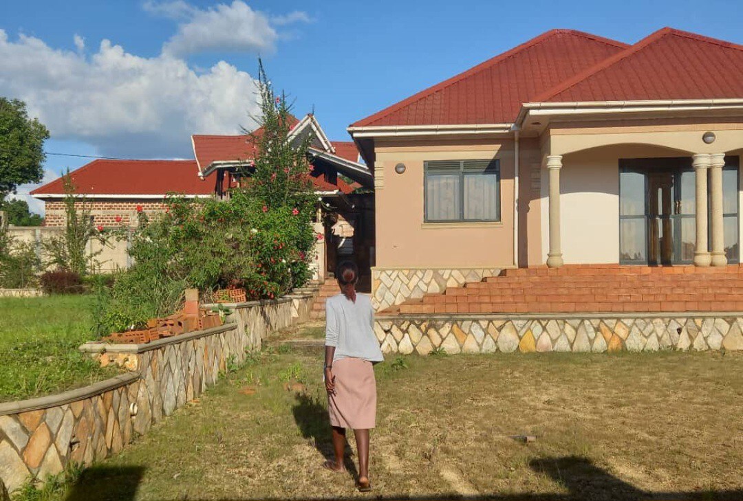 A spacious exterior view of a single-story home is presented, featuring a sloped roof and stone accents. A person is approaching the entrance, which is framed by a set of steps. Lush greenery and a garden area are visible, alongside a blue sky with scattered clouds.
