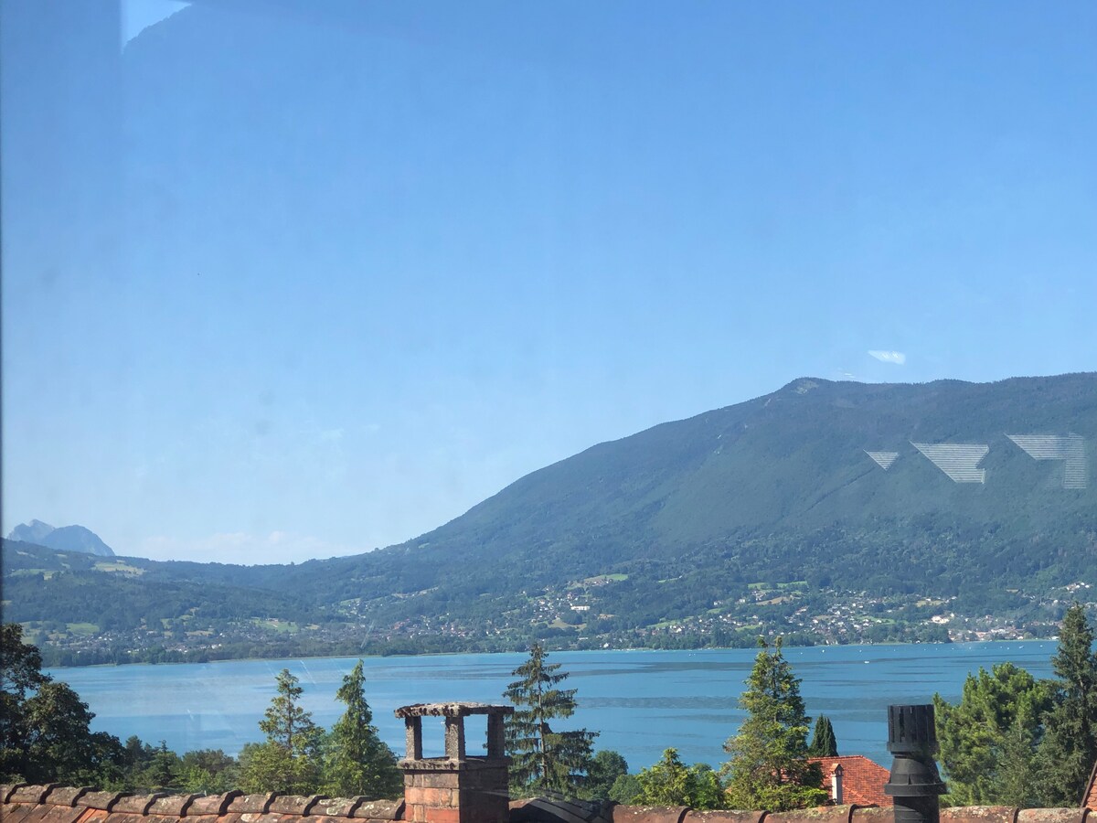 A serene view of the lake is framed by distant mountains and lush greenery. The clarity of the water reflects the surrounding landscape under a clear blue sky, inviting an experience of tranquility and connection with nature.