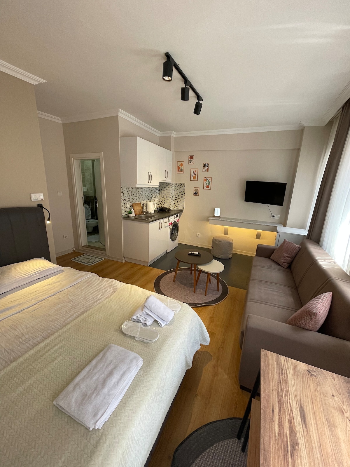 A cozy studio space is furnished with a spacious bed, a comfortable sofa, and a small coffee table. A kitchenette with modern amenities is visible along one wall. Natural light fills the room through large windows, complementing the neutral color palette.