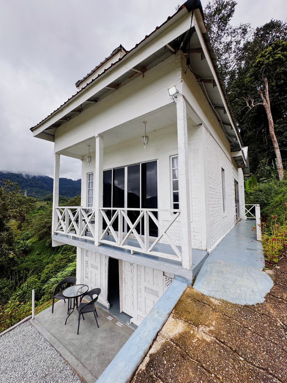 A two-story white cottage is positioned on a hilltop, featuring a broad balcony with two black chairs and a small table. Large glass doors provide access to the interior, while lush greenery and expansive views of the surrounding hills are visible.