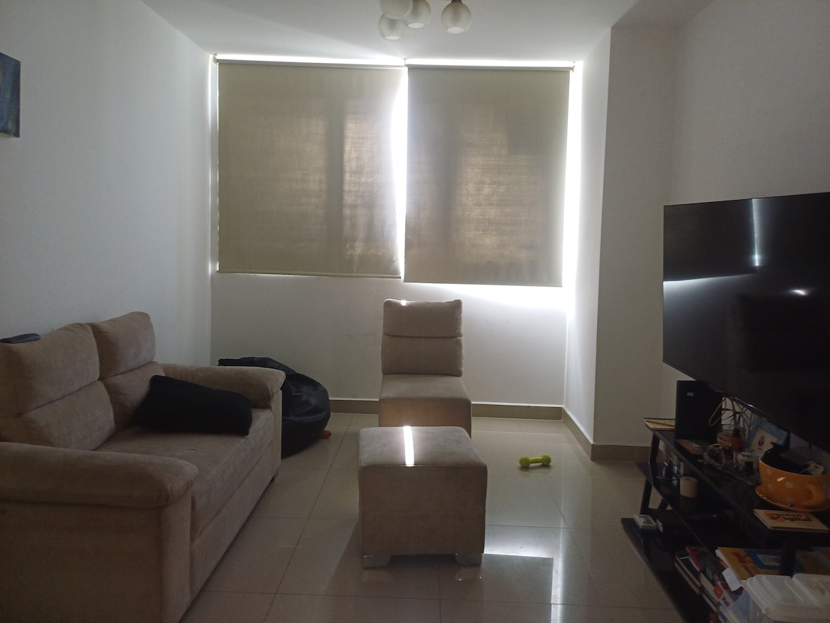 A light-filled living area features a comfortable beige sofa and two upholstered stools. Natural light streams in through partially closed beige blinds. A dark television sits on a low media console, with various items arranged on it. A soft toy lies on the shiny tiled floor.