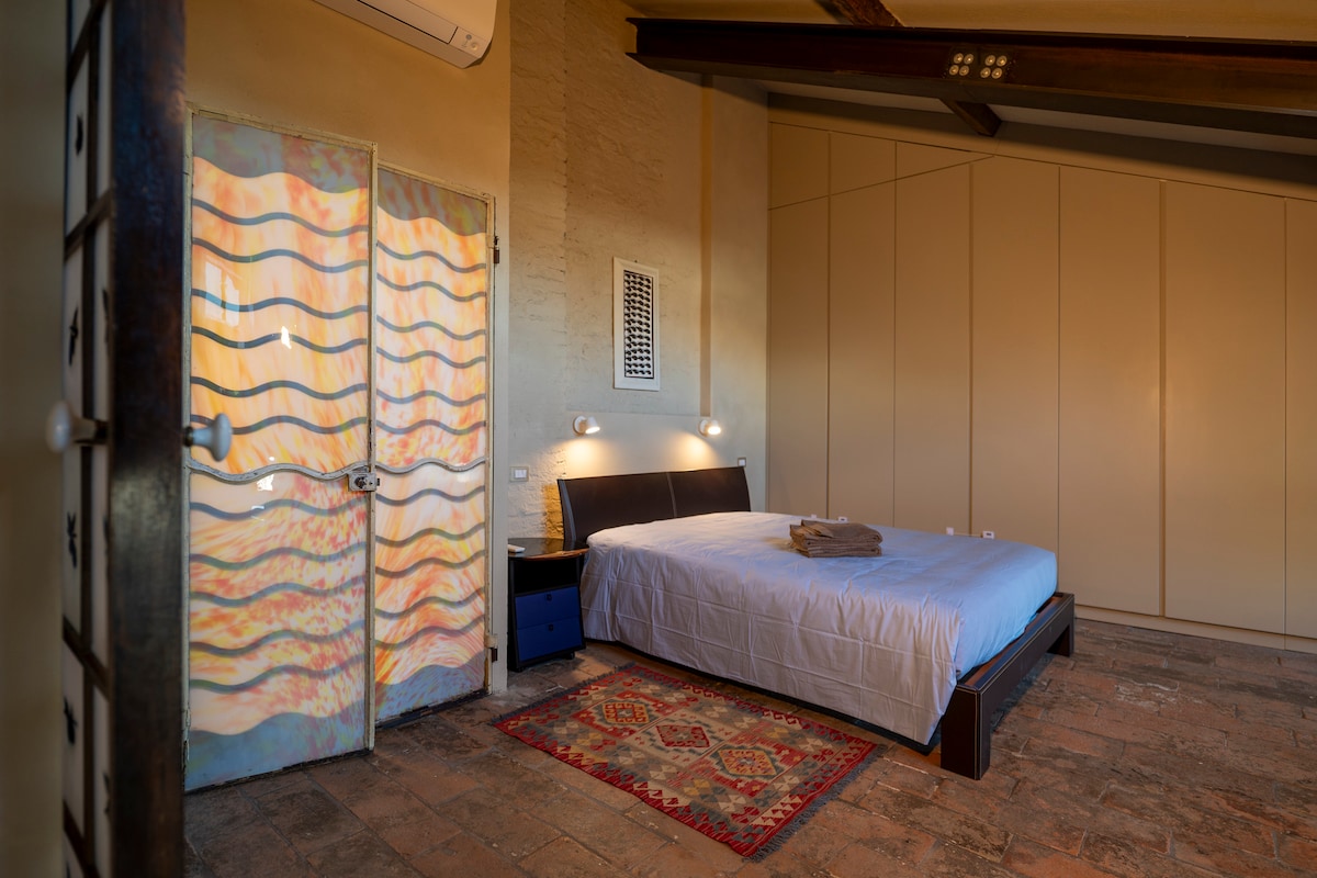 Dormitorio principal de 30 metros cuadrados, con techo muy alto, cama doble Molteni con colchón de muelles de 30 cm, pared con armario de pared equipado, cómoda antigua, equipo de música y acceso directo al baño. Hay una ventana alta y una ventana baja.