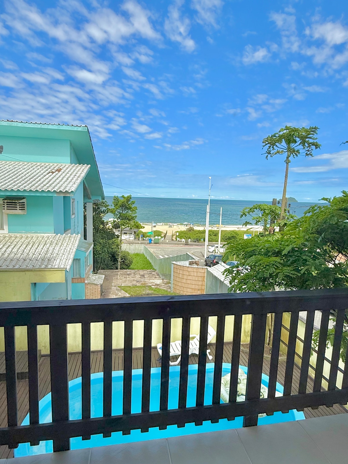 A view from the balcony highlights the serene blue sea and sky. A swimming pool is visible below, surrounded by greenery. The distance showcases a pathway leading to the beach, complementing the coastal atmosphere of the setting.