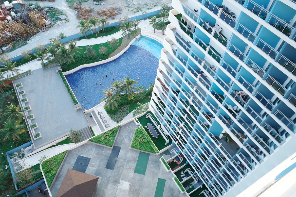 An aerial view captures the expansive pool area surrounded by palm trees, featuring a sparkling blue wave pool. The multi-story building is visible, showcasing multiple balconies. A landscaped outdoor space with seating and grassy areas can also be seen adjacent to the pool.