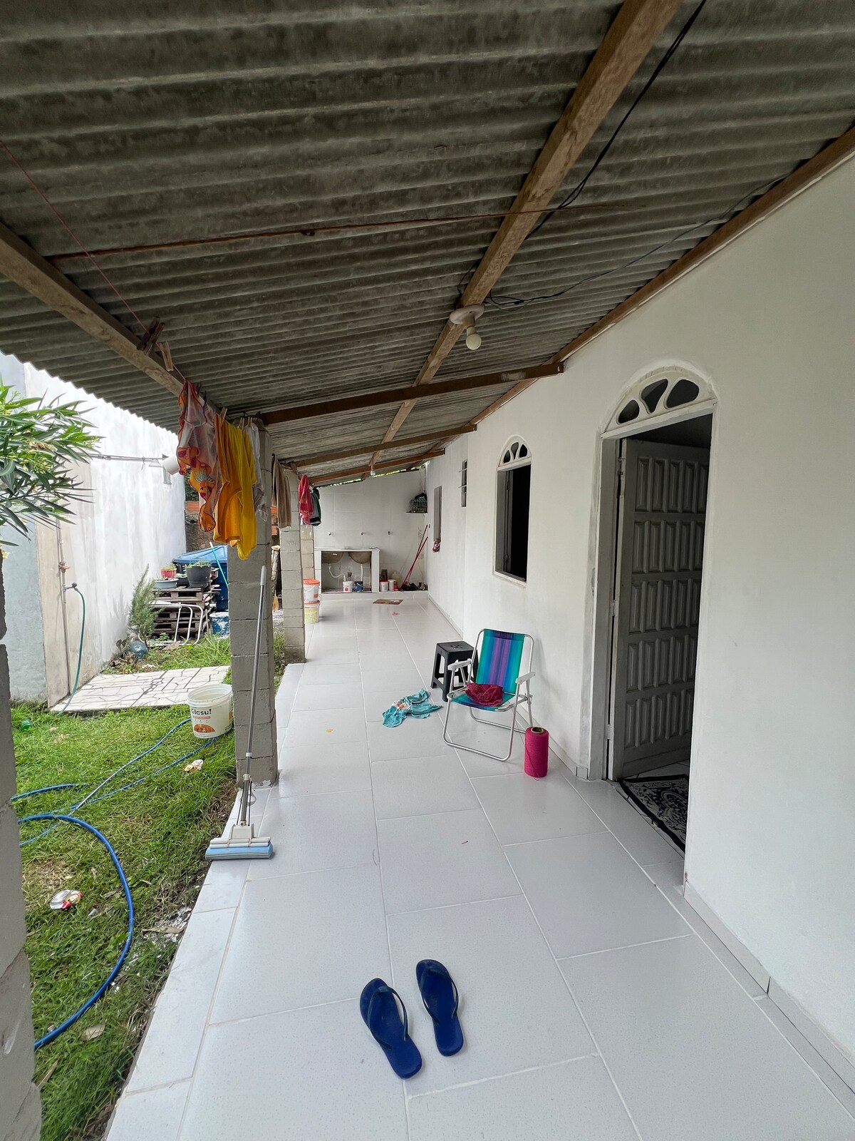 An outdoor corridor is observed, lined with white tiles and supported by a rustic metal roof. Colorful clothing is hung to dry above, while a few chairs and a small table are positioned along the way. Green grass is visible to one side, creating an inviting atmosphere.