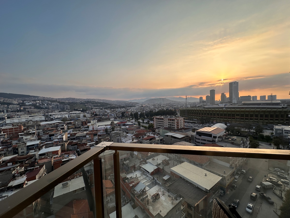 A panoramic view from a high balcony captures the city skyline at sunset. The horizon displays a gradient of warm colors, while buildings and rooftops populate the landscape below. The sleek glass railing offers unobstructed sightlines to the sprawling urban environment.
