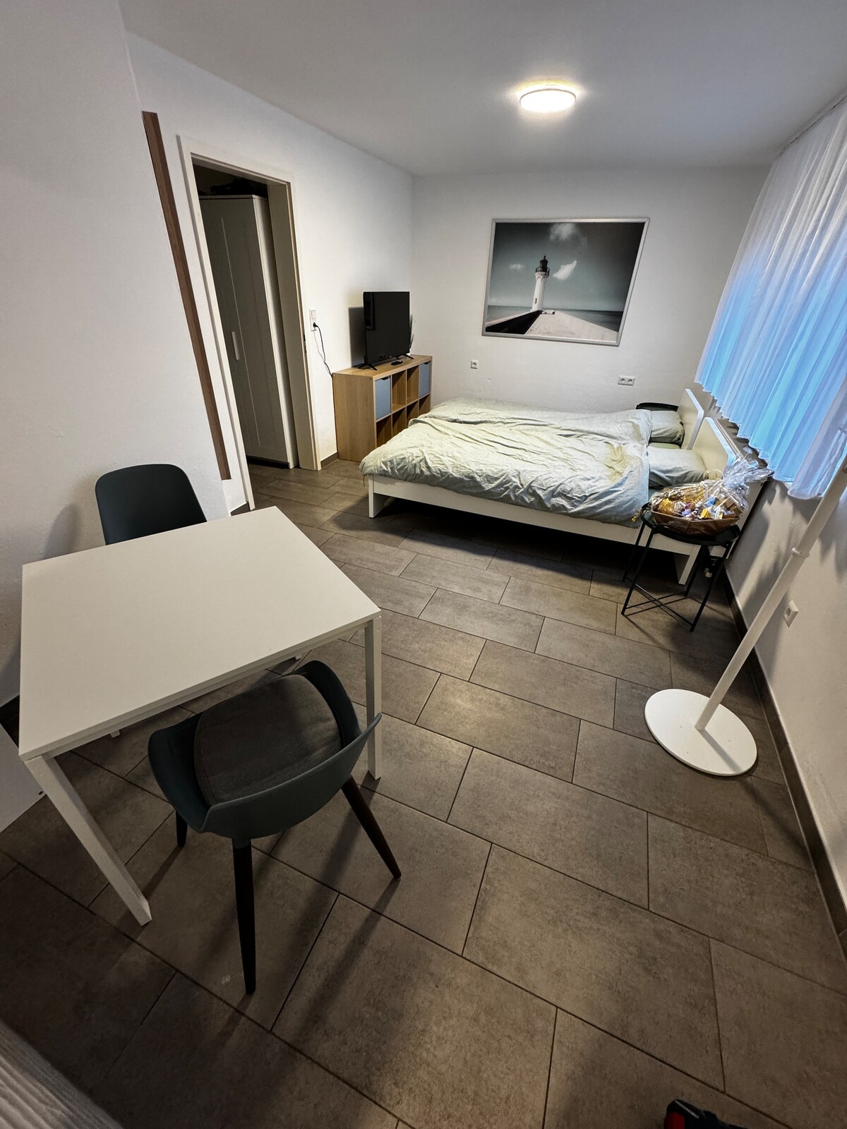 A comfortable living space is presented with two single beds against a wall, complemented by a gray armchair and a small white table. A TV is mounted on the wall near a mirror, with light filtering through sheer white curtains beside a partially open closet.