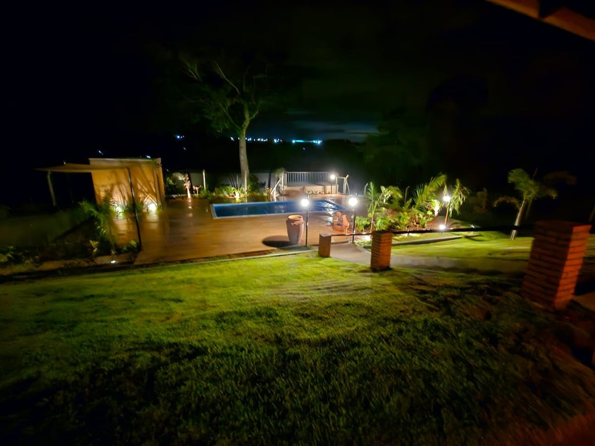 A serene outdoor area is featured with a well-lit pool surrounded by wooden decking and landscaped greenery. Soft lighting accents the space, providing a calming atmosphere. A seating area and patio are visible, creating an inviting spot for relaxation.