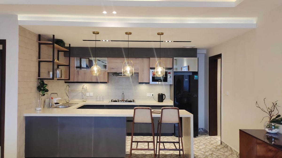 A modern kitchen space features sleek cabinetry in wood and gray tones. Three pendant lights illuminate the countertop. A gentle contrast is provided by two high stools positioned at the breakfast bar. Appliances are neatly integrated into the design, enhancing the overall functionality.