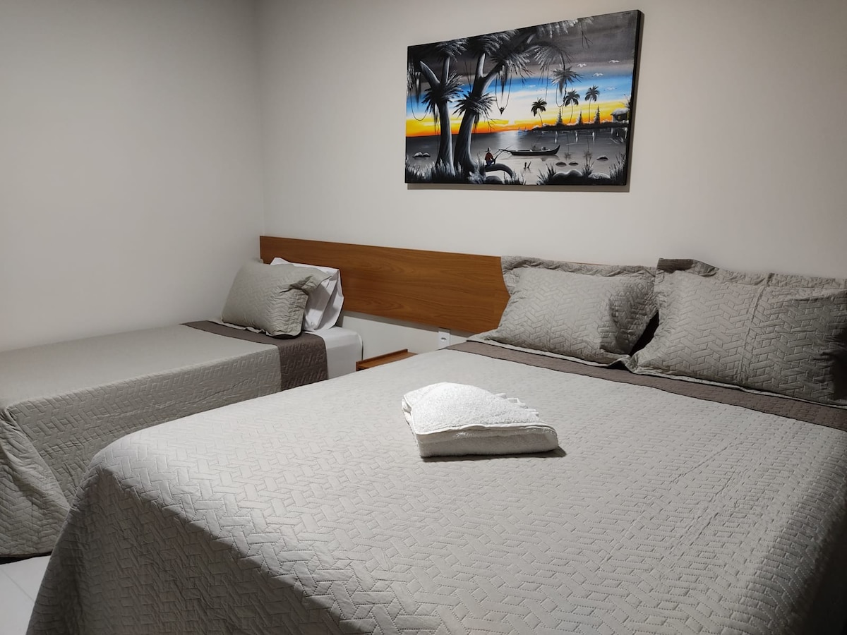 A cozy sleeping area is presented with a spacious bed and a single bed, both covered in light gray bedding. A colorful artwork depicting a sunset scene hangs above the beds, enhancing the overall ambiance of the room.