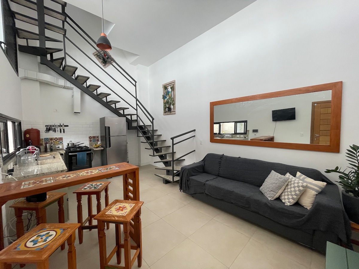 The image depicts a modern open-concept living and dining area with a neutral color scheme. A sleek kitchen features stainless steel appliances and a bar with high stools. A staircase hugs the wall, leading to the upper floors. A comfortable sofa with decorative cushions is positioned beside a large mirror.