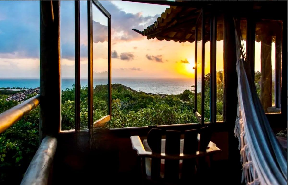 Large windows frame a serene view of the ocean at sunset, with vibrant hues in the sky. A hammock and a wooden chair are positioned on the terrace, surrounded by lush greenery, inviting relaxation and a connection to nature.