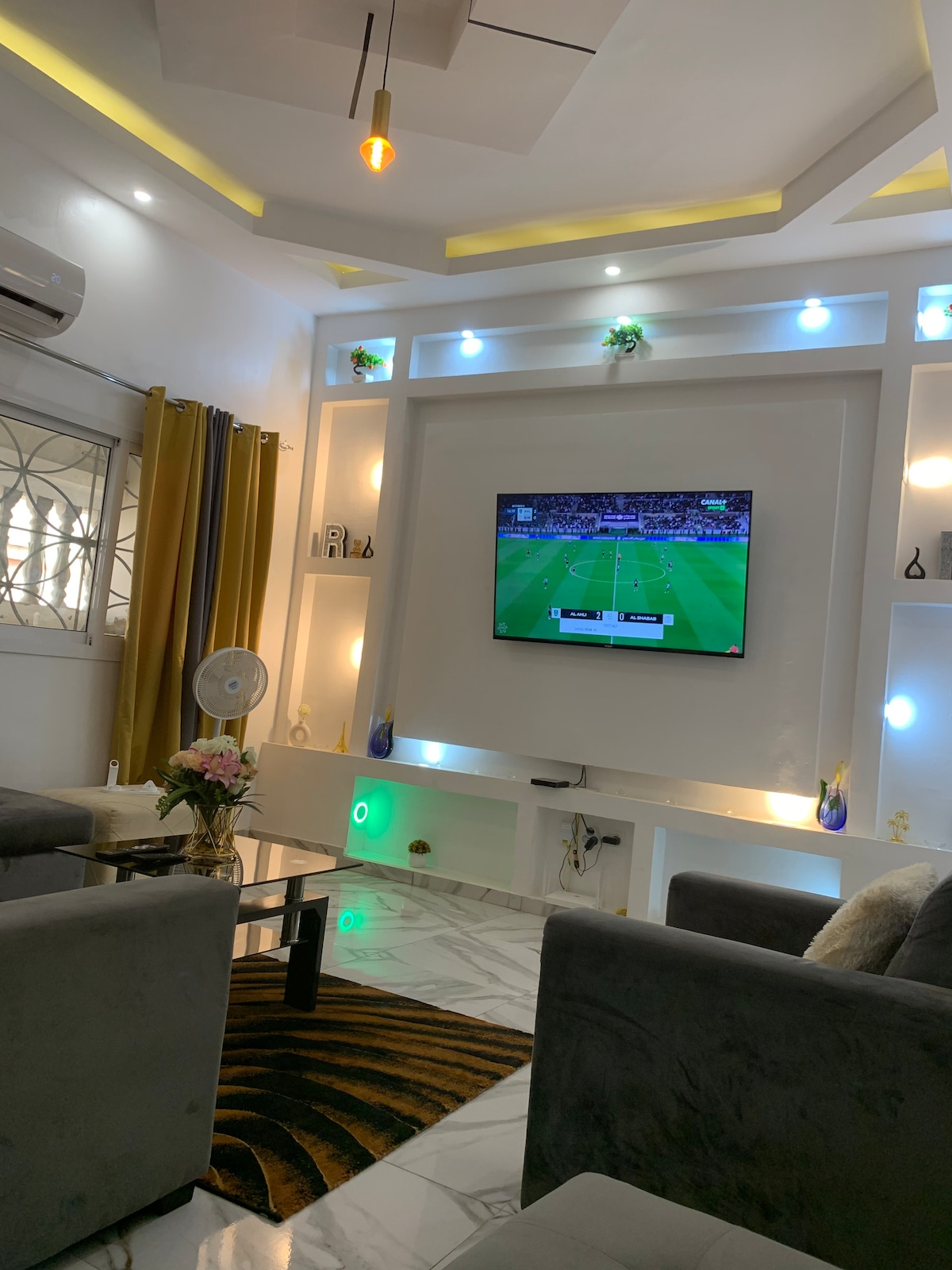 A modern living area features a wall-mounted television displaying a sports game. Soft grey sofas are positioned around a glass table, with decorative lighting in soft hues illuminating the space. A potted plant and a small fan are also visible, enhancing the room's comfort.
