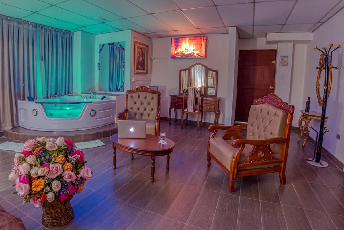 A spacious living area features two ornate armchairs facing a wooden coffee table. A modern television is mounted on the wall above a Jacuzzi tub glowing with ambient blue lighting. A decorative flower arrangement adds a touch of color, complemented by a wooden dresser in the background.