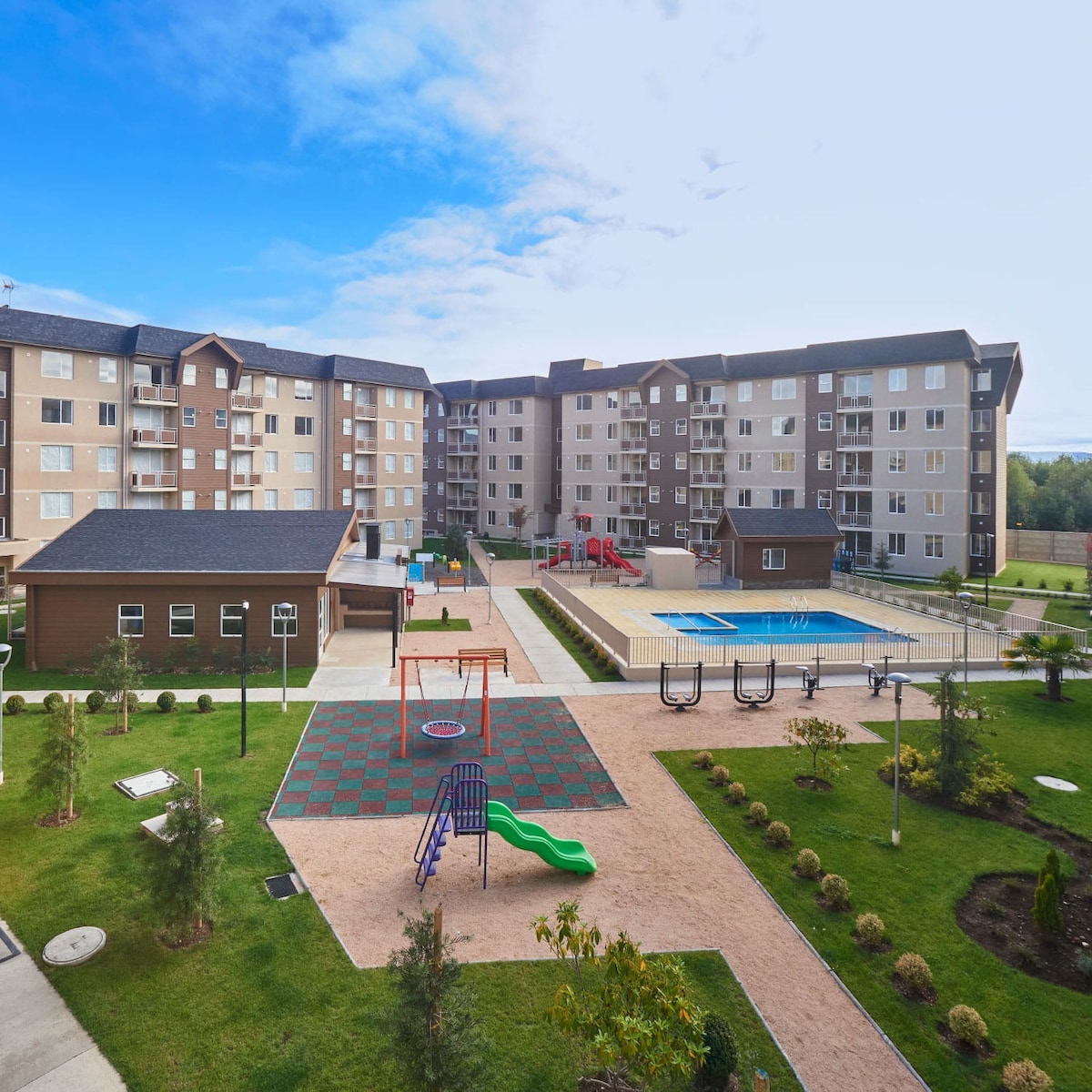 The image depicts a spacious courtyard within a residential complex. A swimming pool is visible alongside grassy areas, playground equipment, and seating. The buildings are arranged harmoniously, providing a welcoming outdoor space for relaxation and recreation.