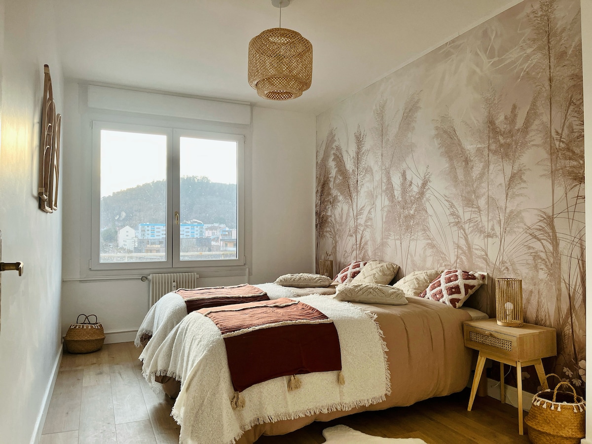 A serene bedroom features a spacious bed adorned with soft textiles in earthy tones. Natural light fills the room through large windows, offering a view of the surrounding hills. Decorative elements include a woven pendant light and a bedside table with minimalistic decor.
