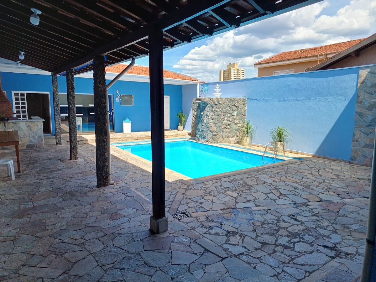 A spacious outdoor area features a rectangular pool surrounded by stone flooring. Tall wooden beams support the roof, providing shade. The walls are painted in shades of blue, and the pool is complemented by subtle landscaping elements, creating a relaxed and inviting environment.