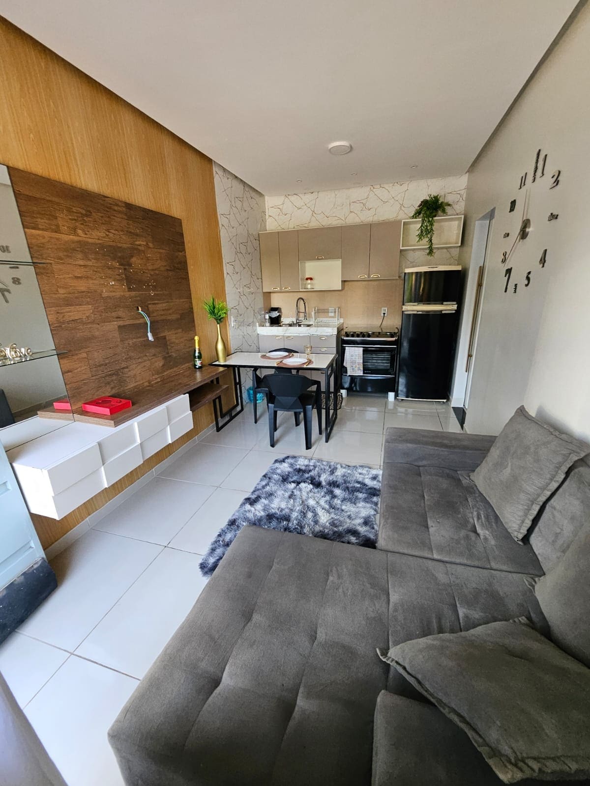 A modern living area features a gray sofa and a dining table set for four. The compact kitchen is visible, with a refrigerator and stove, surrounded by light wood and patterned tile accents. Natural light fills the space through a window, enhancing the contemporary design.