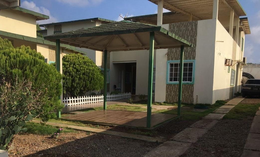 An open patio area is featured under a shaded gazebo, surrounded by well-maintained greenery and a white picket fence. The adjacent pathway leads to the entrance of the property, which is flanked by two light-colored structures.