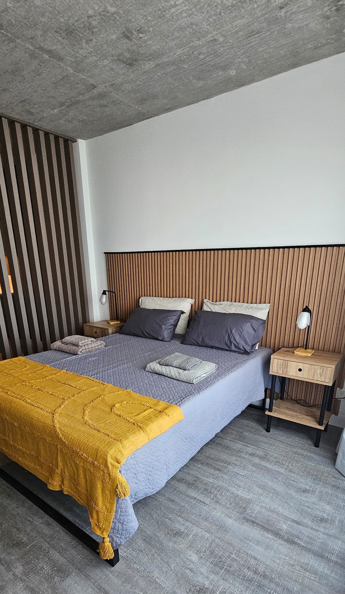 A modern sleeping area features a comfortable queen-size bed with a light quilted cover and pillows. Wooden side tables are positioned on either side, showcasing simple lamps. A textured accent wall adds depth, while large windows allow natural light to illuminate the space.