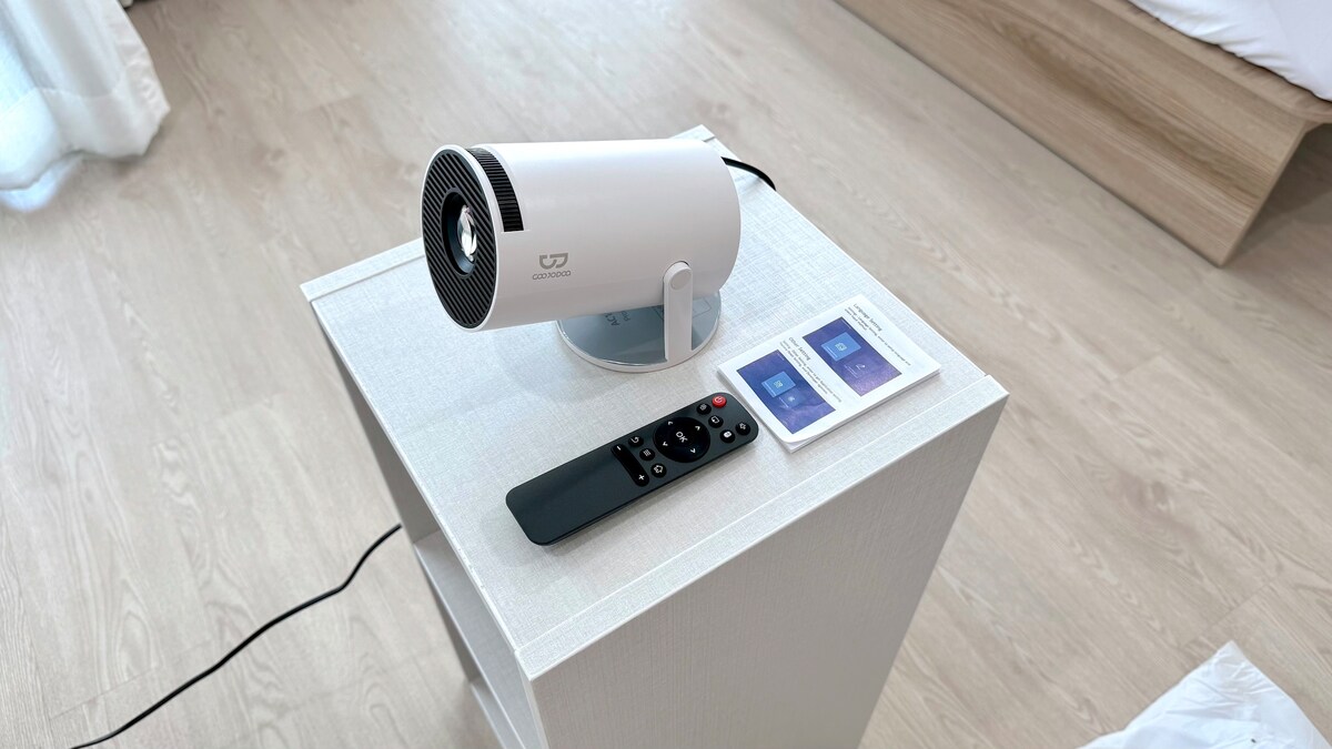 A compact air purifier is positioned on a sleek white bedside table. The remote control and user manual are placed alongside it, emphasizing convenience. The light-colored floor and soft materials in the background provide a calm setting.