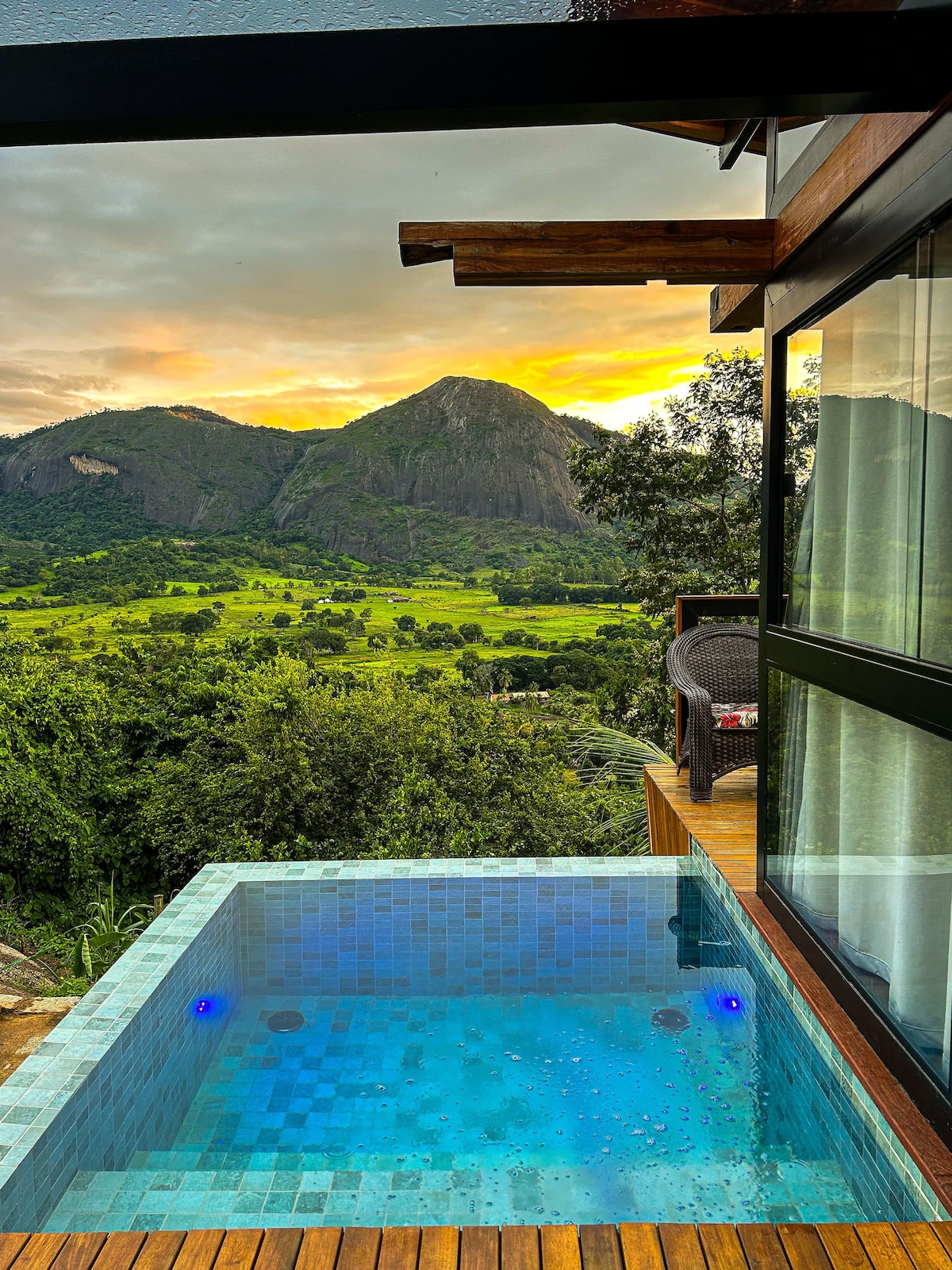 A private hot tub is positioned on a wooden deck, overlooking an expansive valley framed by mountains. The sunset casts warm hues across the sky, while the glass walls of the chalé reflect the natural beauty surrounding the space.