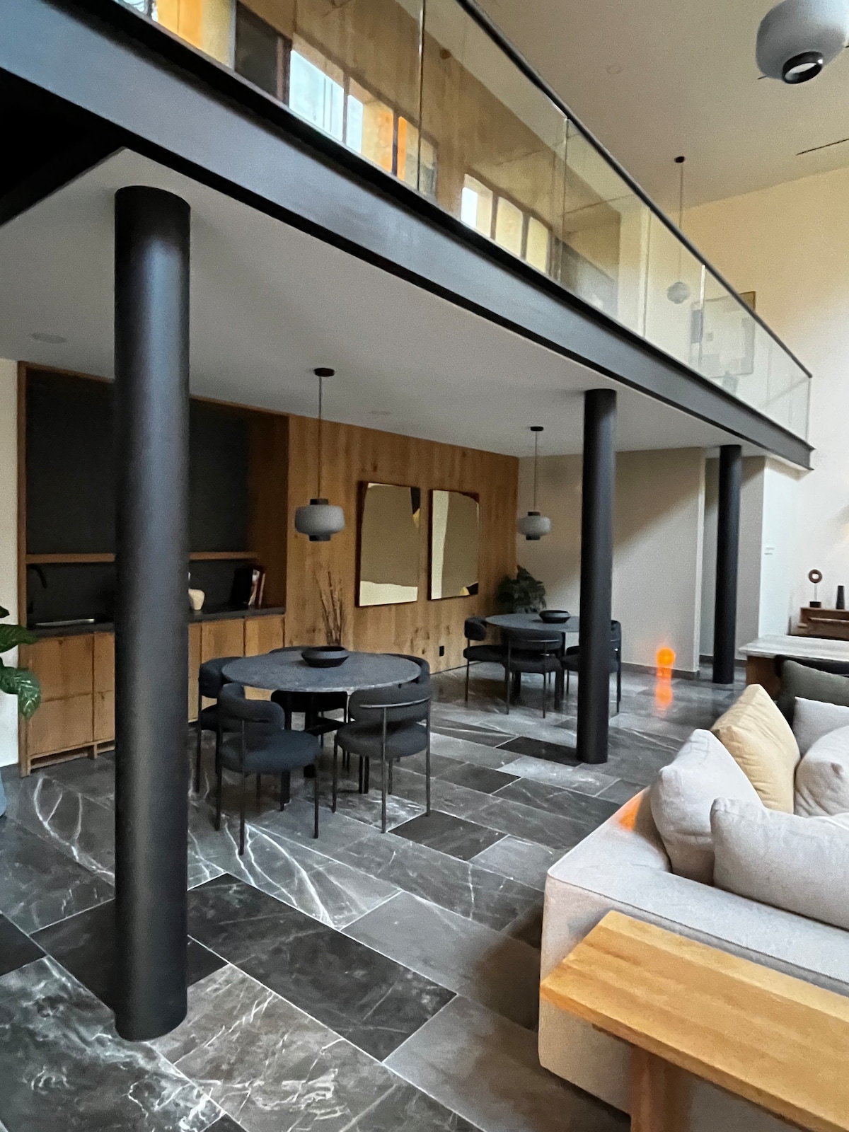 An open dining area features a round black table with black chairs surrounded by elegant wooden accents. A spacious living area showcases a light sofa and large windows, allowing natural light to fill the space. Contemporary lighting fixtures hang above, complementing the harmonious design.