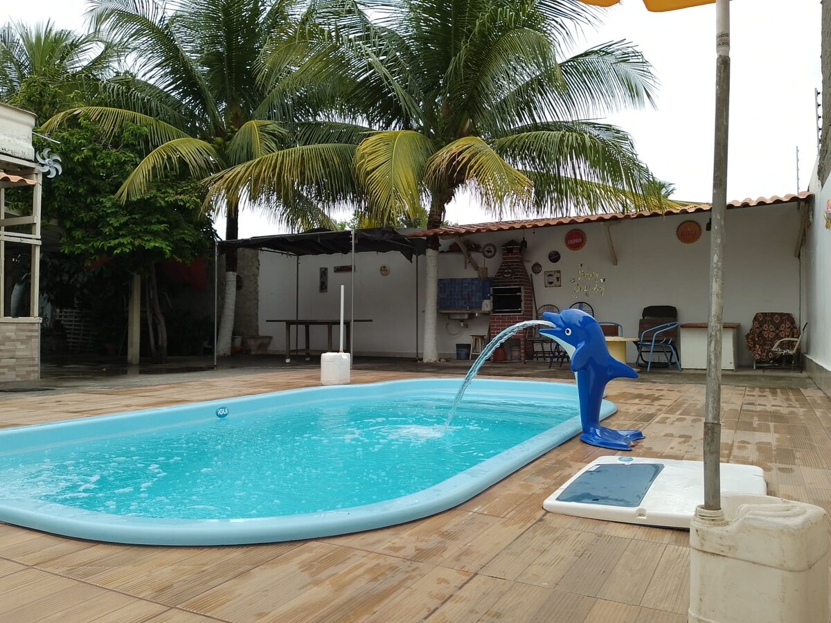 A swimming pool is surrounded by wooden decking and tropical palm trees. A playful dolphin-shaped water feature adds charm, while the area includes a shaded seating space. A partially covered patio provides additional relaxation options in a spacious outdoor environment.