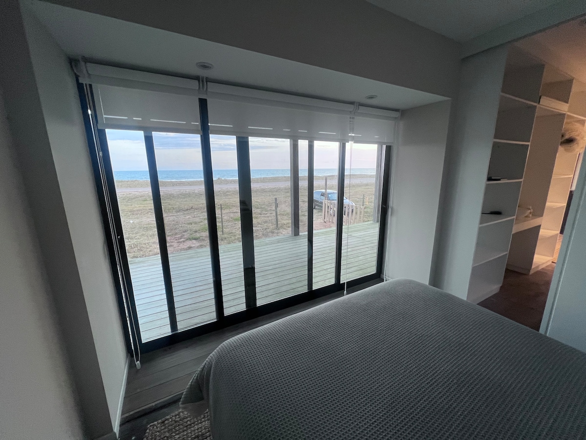 A bright bedroom features expansive glass doors that provide a clear view of the beach and sea. Natural light fills the space, while a neutral-colored bedding complements the minimalistic decor, creating a calming atmosphere for relaxation.