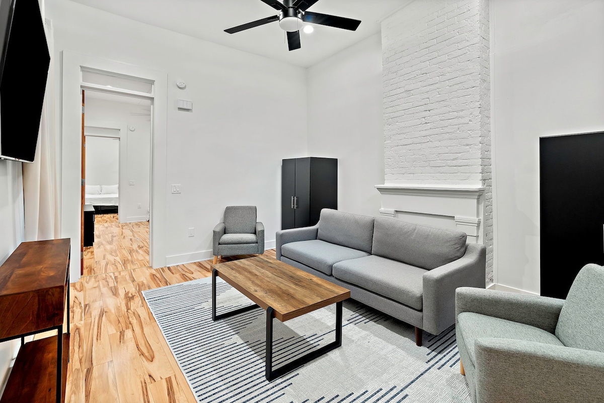 The living area features a light-colored sofa and two chairs arranged around a wooden coffee table. A textured area rug lies beneath, while a large television wall mount is visible. The exposed brick wall adds an element of texture to the bright, inviting space.